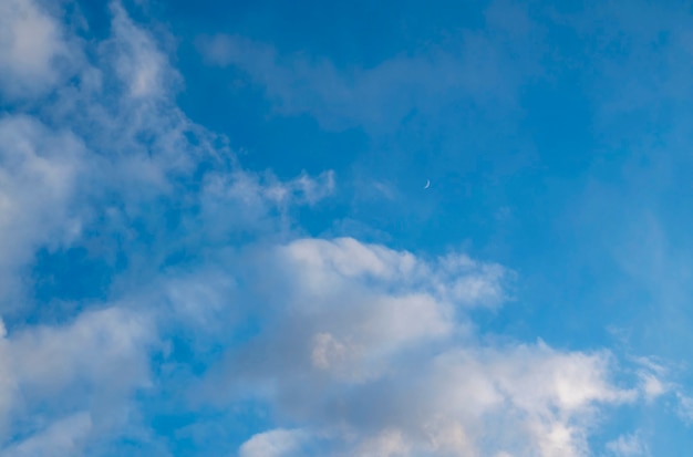 Nuvens em um céu azul com uma lua crescente à distância ao pôr do sol.