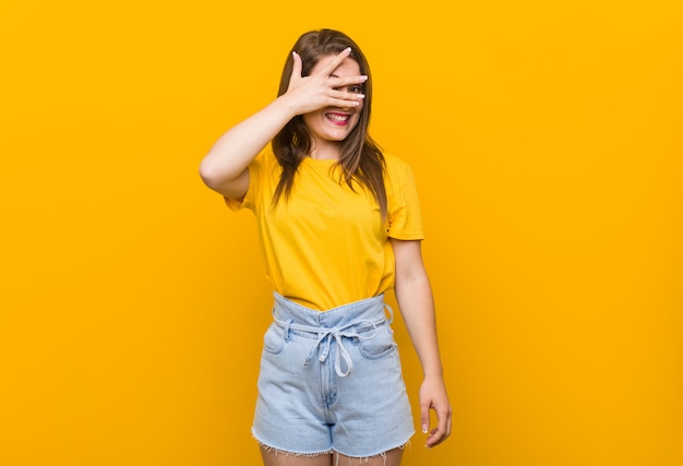 O adolescente da jovem mulher que veste uma camisa amarela pisca através dos dedos, cara de coberta embaraçado.