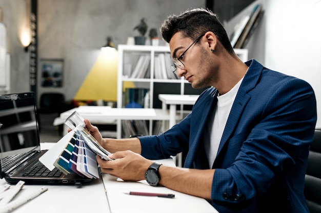 O arquiteto de óculos e de paletó azul está trabalhando com catálogo de cores na mesa do escritório.