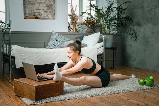 O atleta se alongando, meditando, sentado em uma corda ee assiste no laptop, fotografando o blog no quarto, ao fundo tem uma cama, um vaso, um tapete.