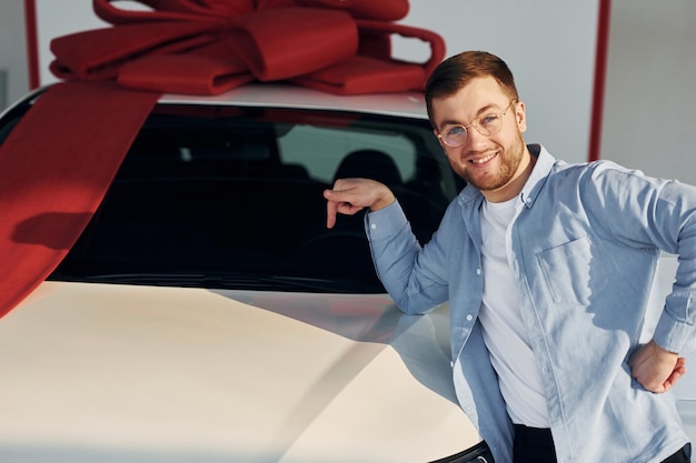 Foto o automóvel está coberto de fita de presente homem bem sucedido em óculos parado perto de um carro novo dentro de casa