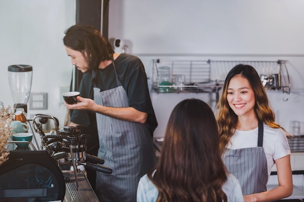 Foto o avental asiático bonito novo do desgaste do barista da mulher que guarda o copo de café quente serviu ao cliente no contador da barra na cafetaria com cara do sorriso.