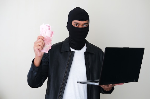 Foto o bandido usando uma balaclava roubando dinheiro usando um laptop