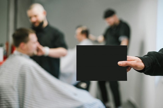 O barbeiro tem um cartão em branco preto na mão para o espaço da cópia no fundo dos barbeiros que cortam clientes. Masculino mão segurando um cartão em branco sobre fundo de barbearia.