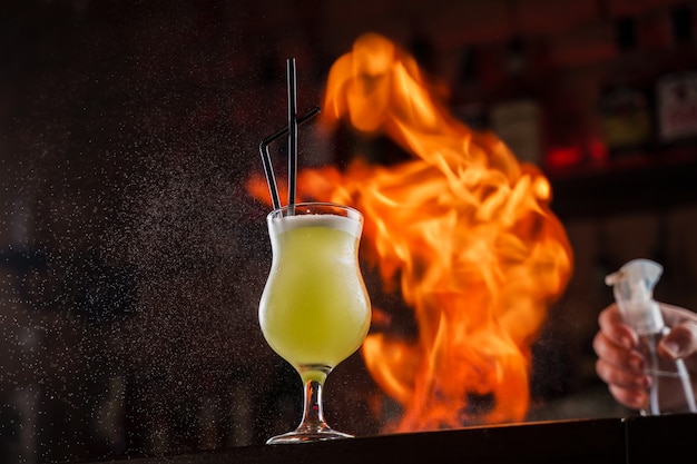 O barman polvilha no vidro iluminado com um coquetel frio verde brilhante no balcão do bar e faz o fogo acender sobre ele.