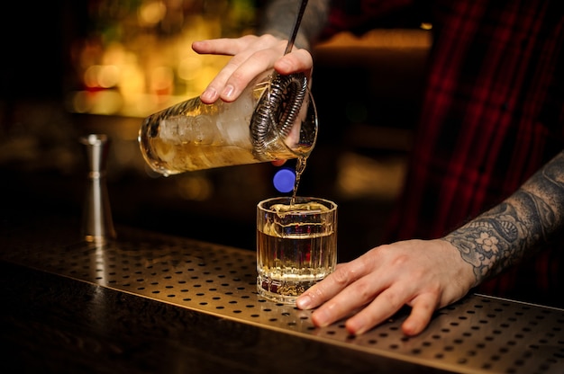 O barman servindo um coquetel de unhas enferrujadas do copo medidor através da peneira em um copo no balcão do bar