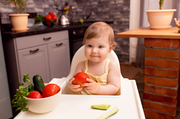 O bebê alegre 10-12 meses come vegetais. Retrato de uma menina feliz em uma cadeira na cozinha