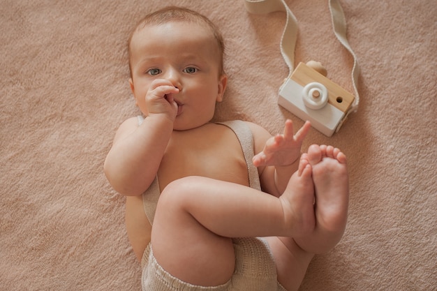 Foto o bebê com uma câmera de madeira deitado sobre um fundo bege, o bebê chupa o dedo