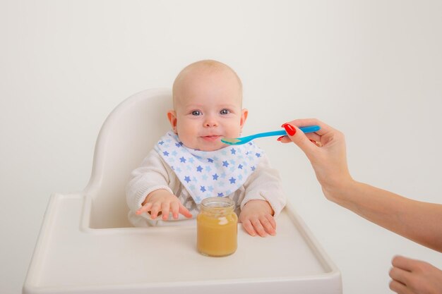 O bebê está sentado em uma cadeira alta a mãe alimenta o bebê com purê de frutas em um fundo branco