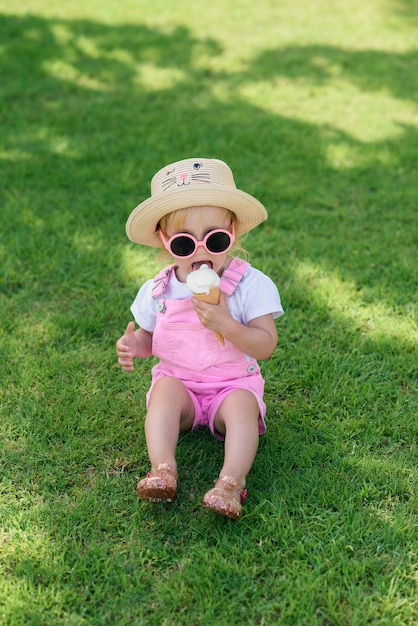 Foto o bebê feliz vestiu a roupa cor-de-rosa do verão, chapéu amarelo e óculos de sol cor-de-rosa senta-se em um gramado verde e come-se o gelado branco em um jardim ensolarado.