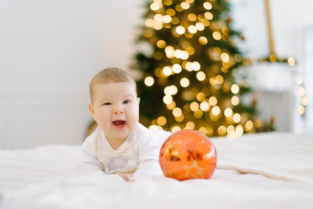 O bebê ri, cobrindo um olho, deitado na cama contra o fundo de luzes de Natal