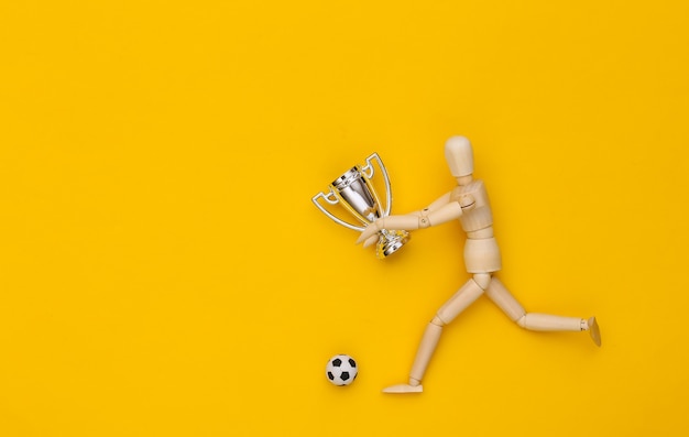 O boneco de madeira corre com a bola de futebol e a taça do campeonato em fundo amarelo