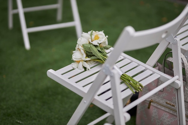 O buquê de casamento está em uma cadeira branca dobrável no contexto de um gramado cortado