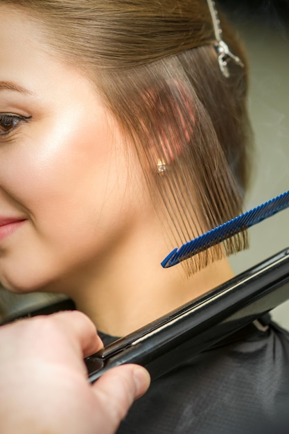 O cabeleireiro está endireitando o cabelo curto da jovem morena com uma chapinha