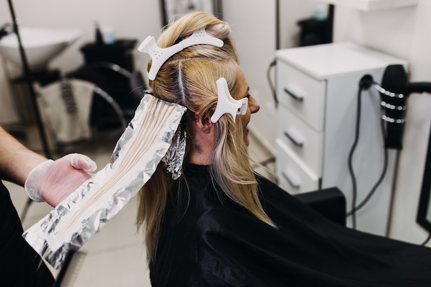 O cabeleireiro está tingindo o cabelo feminino, fazendo mechas de cabelo para sua cliente com um papel alumínio.