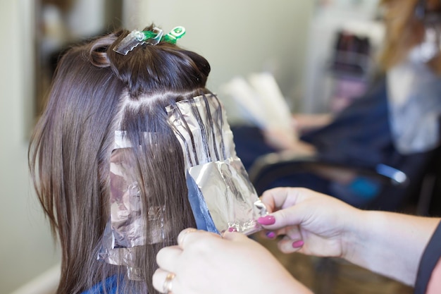 O cabeleireiro pinta o cabelo de uma mulher morena no salão de beleza perto