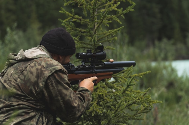 O caçador mira com um rifle. Um homem camuflado está se preparando para atirar. Caçando na floresta com um rifle de precisão