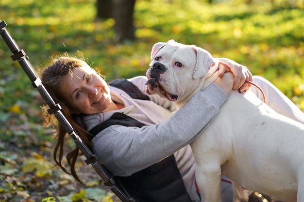 O cachorro brinca com a amante no parque Closeup de uma mulher em uma jaqueta e um cachorro bulldog americano brincando entre as folhas amarelas de outono no parque