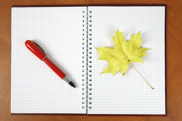 Foto o caderno aberto e a caneta vermelha com folhas de outono sobre uma mesa de madeira