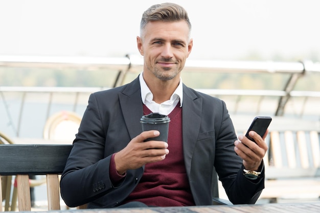 O café me completa O chefe segura a xícara para viagem e o smartphone Pausa para o café ao ar livre Bebida quente Tecnologia moderna Café da manhã da manhã Comunicação empresarial