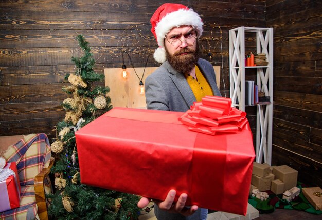 O cara barbudo com óculos carrega a caixa de presente. Presente de Natal de entrega. Serviço de entrega. O Natal está chegando. Entrega de presentes. Correio do Papai Noel. Chapéu de Papai Noel homem entregar presente. Espalhe felicidade e alegria.