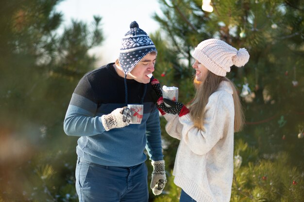 O cara com a garota anda e beija na floresta de inverno com uma caneca de bebida quente