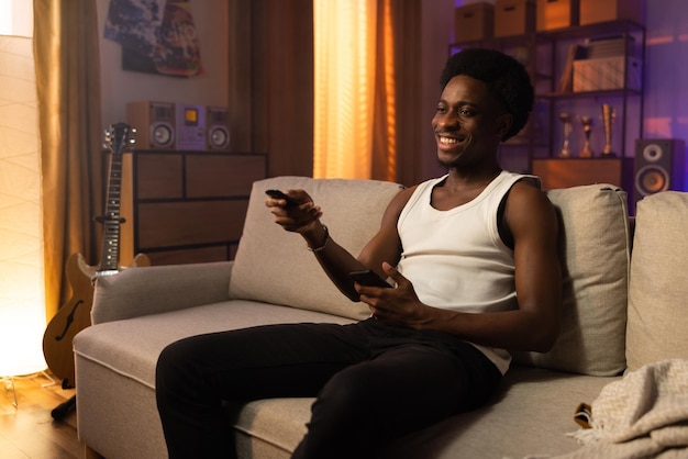 O cara descontraído relaxa no sofá na sala de estar do homem afro em uma camiseta branca sem alças segura o telefone