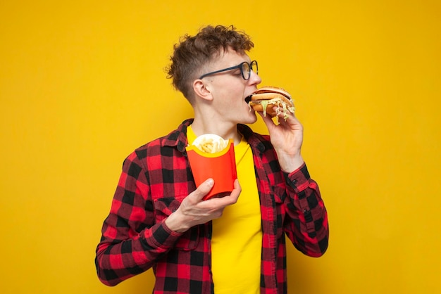 O cara encaracolado segura batatas fritas e morde um grande e saboroso hambúrguer em um estudante de fundo amarelo que come fast-food