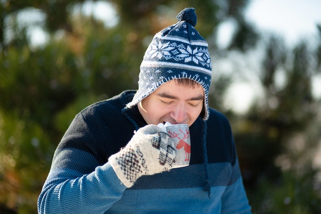 O cara está bebendo uma bebida quente com marshmallows no inverno na floresta. um inverno aconchegante caminhar pela floresta com uma bebida quente. há marshmallow de uma caneca com uma bebida. foto engraçada