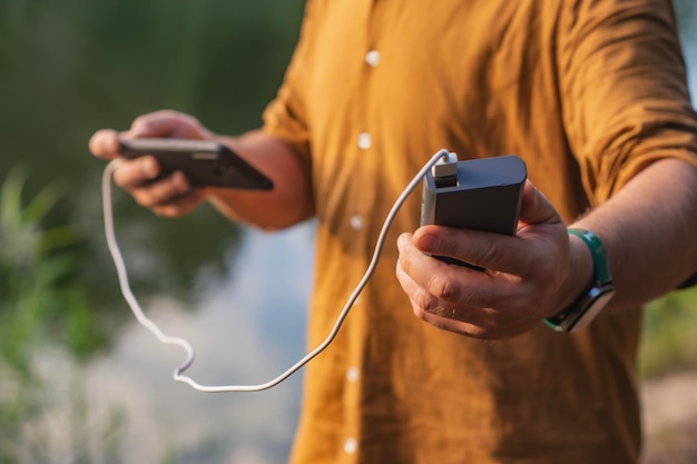 O cara está segurando um carregador portátil com um smartphone na mão Homem em um fundo de natureza com vegetação e um lago