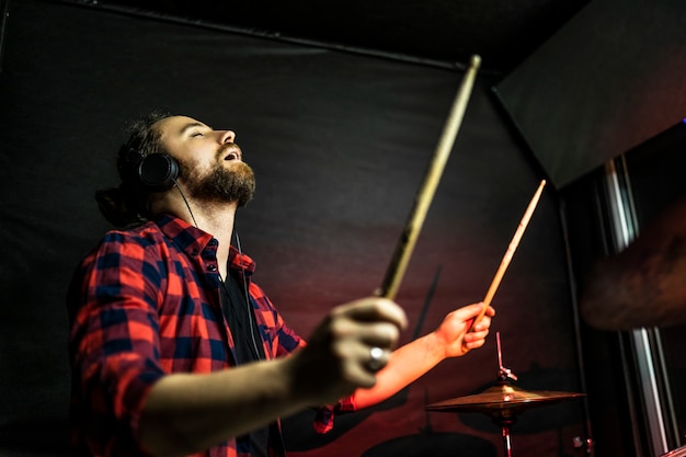 O cara hipster com barba em fones de ouvido está tocando bateria e pecando no estúdio estéreo