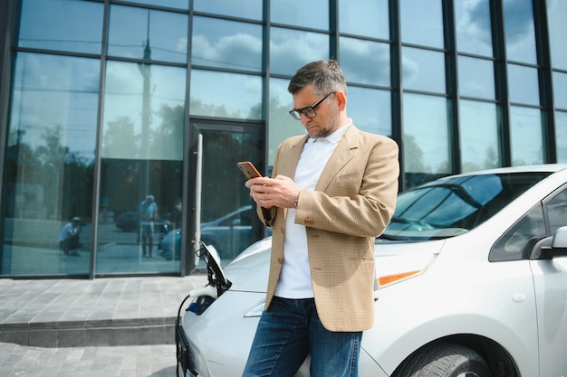 O cara sentou-se no capô do carro Seu carro está carregando na estação de carregamento Um homem olha para a tela do smartphone e sorri