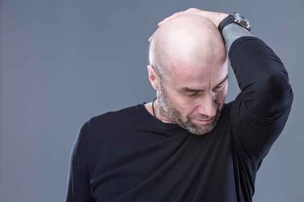 O careca pensativo abaixou a cabeça Um cara brutal com barba por fazer em uma camisa preta segura a cabeça com a mão Fundo cinza Espaço para texto