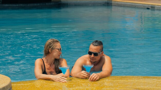 O casal amoroso abraça e beija bebendo licor de álcool coquetel azul na piscina do hotel ao ar livre retrato de homem e mulher caucasianos penteados criativos maiô de fisiculturista óculos de sol