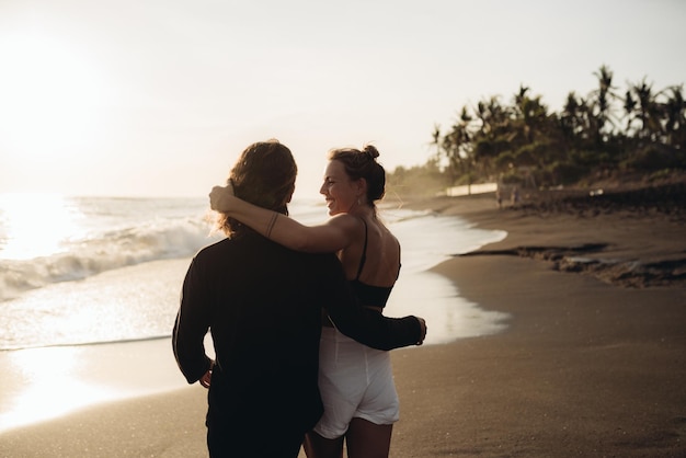 O casal apaixonado filmado pelas costas em um abraço fica na areia costeira Homem abraça mulher pela cintura e olha para as águas do oceano enquanto ela tece suavemente o cabelo na parte de trás de sua cabeça