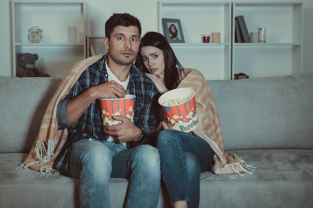 Foto o casal assustado comendo pipoca e assistindo a um filme de terror no sofá