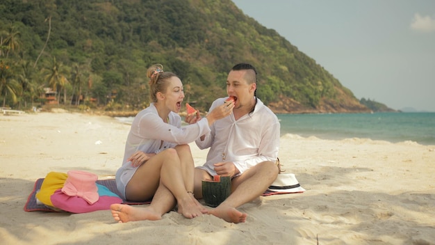 O casal de amor alegre segurando e comendo fatias de melancia no mar de praia de areia tropical romântico