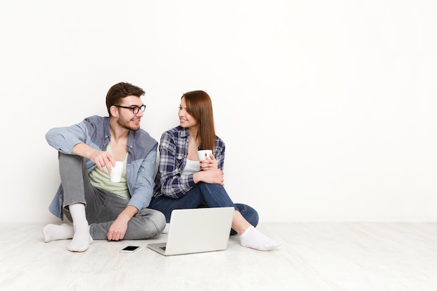 O casal discute algo com o laptop e o celular ao lado deles. homem e mulher felizes sentados no chão no interior branco de seu novo apartamento