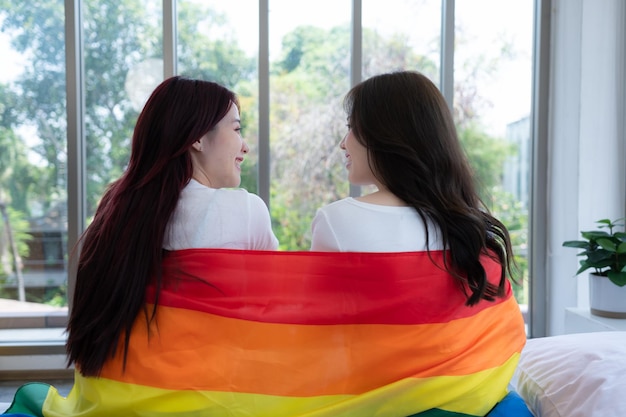 O casal LGBT sentou-se na cama coberto de bandeiras arco-íris olhando pela janela para observar a natureza