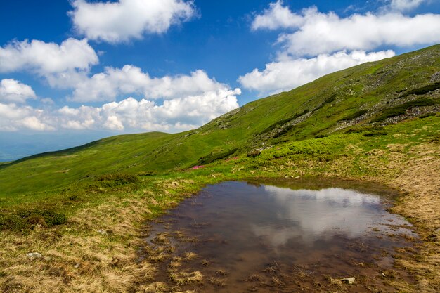 O céu azul com as nuvens brancas brilhantes refletiu no lago pequeno entre montes verdes no dia ensolarado. Panorama de verão.