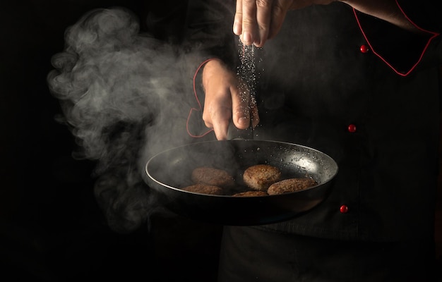 O chef adiciona sal a uma panela quente com costeletas de hambúrguer de carne Espaço livre para menu de hotel ou receita em fundo preto