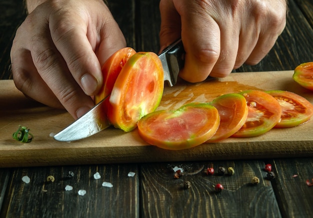 Foto o chef corta tomates em uma tábua de cortar com uma faca com as mãos e uma faca cozinhando salada de legumes para o almoço