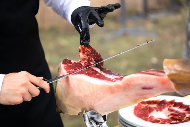 Foto o chef está cortando o presunto. imagem de um cortador de presunto com uma faca cortando uma fatia fina de presunto. presunto seco, presunto serrano, bolota, presunto ibérico, italiano ou parma cru