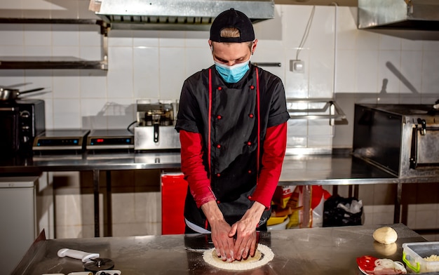 O chef ou fabricante de pizzas, com uma máscara médica contra o coronavírus, faz a massa de pizza em branco. Conceito de fazer massa de pizza