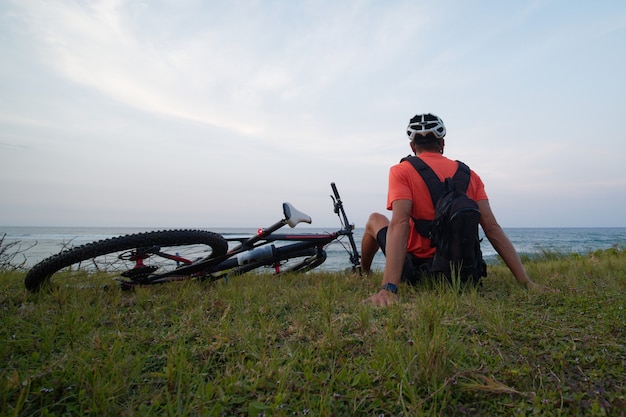 O ciclista com mochila, aproveita a vista para o mar, senta na grama com uma bicicleta. Vista traseira.
