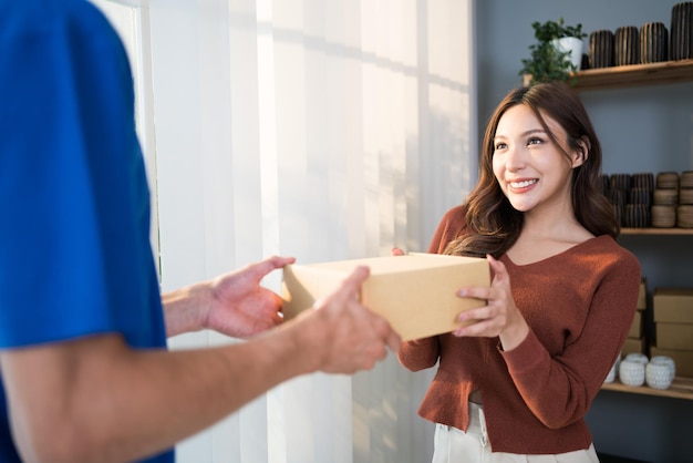 O cliente da mulher asiática recebeu um pacote de envio de encomendas de um mensageiro homem caucasiano em uniforme de mãos dadas na frente da porta de entrada em casa Conceito de serviço de entrega de envio de encomendas em casa