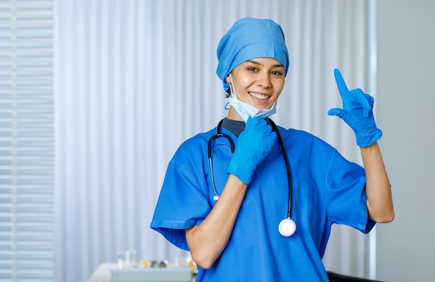 O close up do retrato disparou do feliz muito feminino médico da liberdade em um terno azul do hospital com o estetoscópio sorrindo apontando o dedo para cima quando tirar a máscara cirúrgica usada olhar para a câmera enquanto pandemia termina