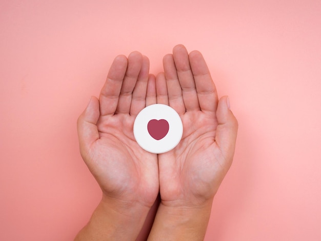 O conceito de amor, cuidado, doação, bem-estar e conceito de doação, estilo minimalista. Mãos segurando o símbolo do ícone de um coração em uma esponja redonda branca sobre fundo rosa pastel.