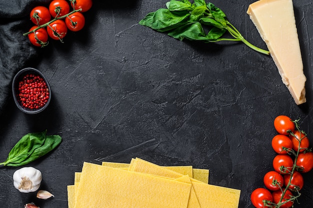 O conceito de cozinhar lasanha. Ingredientes, folhas de lasanha, manjericão, tomate cereja, parmesão, alho, pimenta. Fundo preto. Vista do topo. Espaço para texto