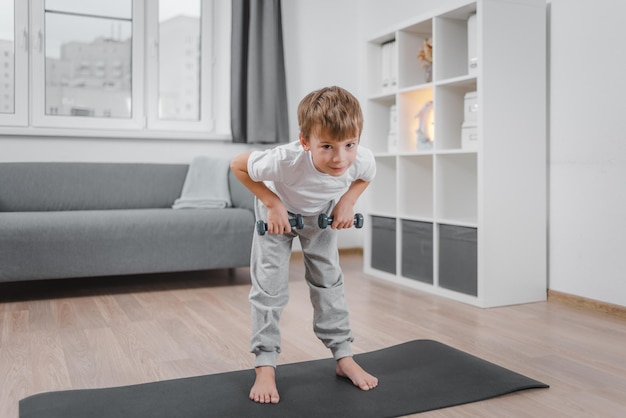 O conceito de esportes durante a quarentena, bloqueio. retrato de menino criança caucasiano fazendo fitness com halteres em casa, na sala de estar.